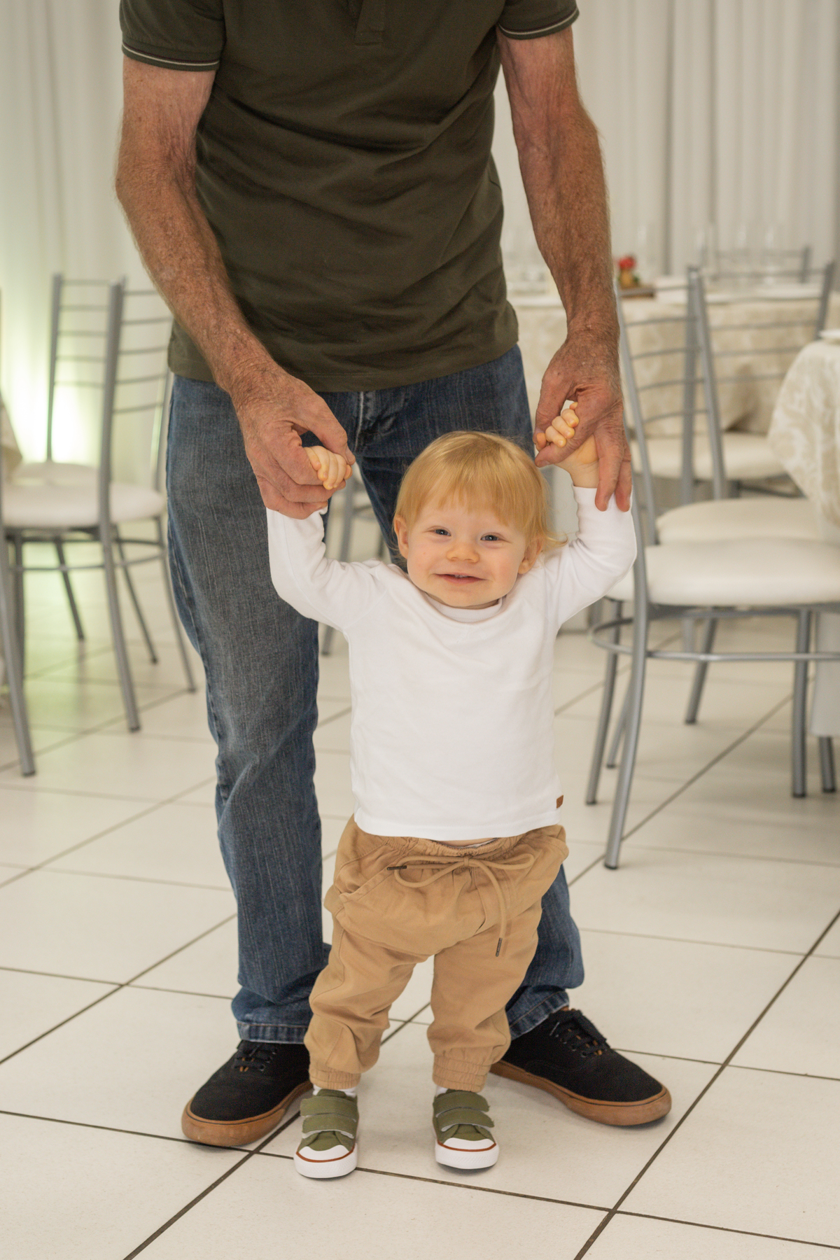 Foto da aniversariante caminhando em direção a fotógrafa com a ajuda do avô, o pequeno sorri.