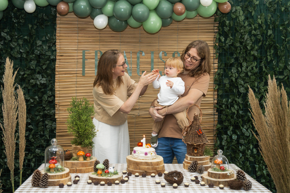 Foto do aniversariante no colo do pai e olhando para a mãe, que bate palmas para ele.