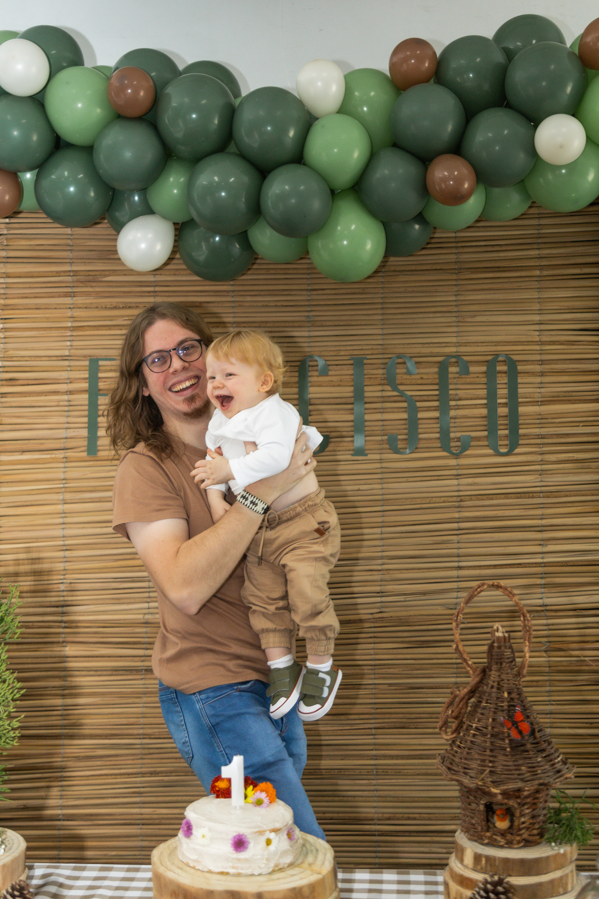 Aniversariante no colo do pai em frente ao painel da festa, ambos sorriem.