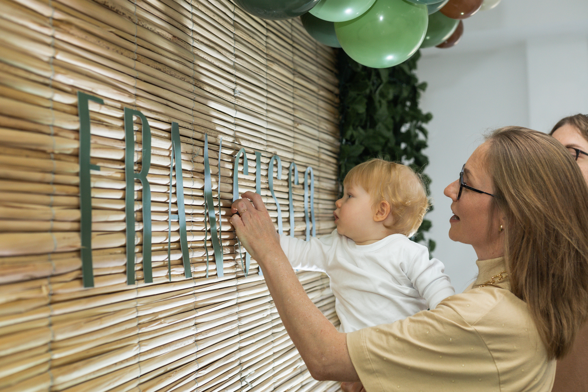 Foto do aniversariante no colo da mãe, observando seu nome escrito no painel da decoração.