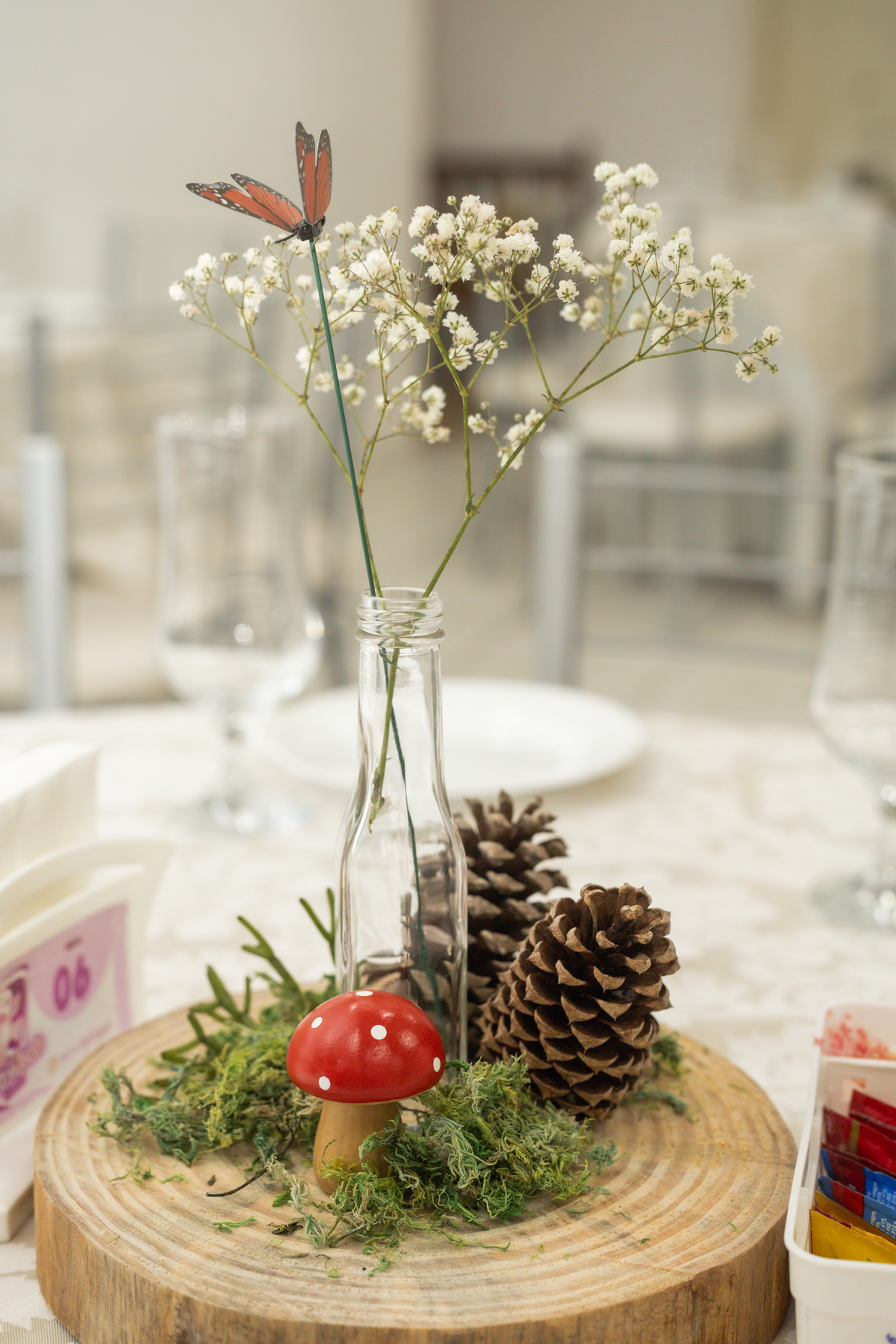 Foto de detalhe da decoração com um centro de mesa de madeira com pinhas, musgo, cogumelo e um vaso com uma flor branca delicada.
