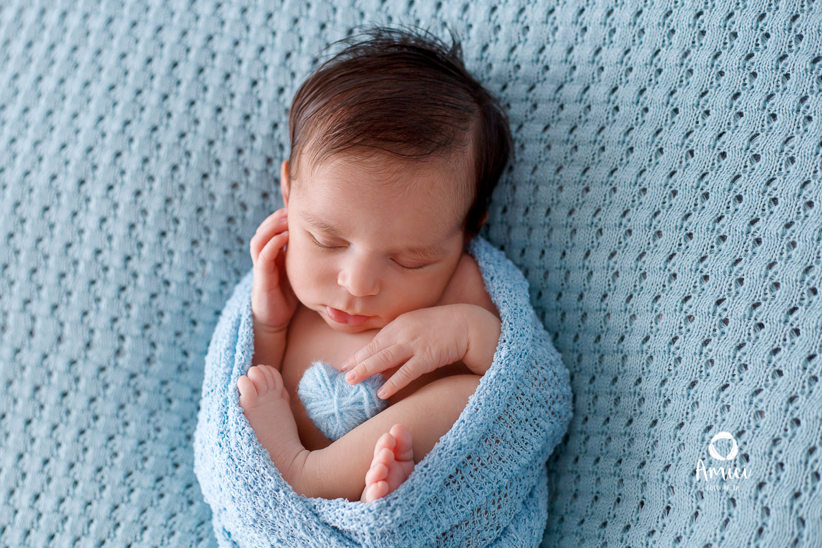 Fotografia newborn, bebê envolvido em um wrap na posição de barriguinha pra cima, segurando um pequeno coração.