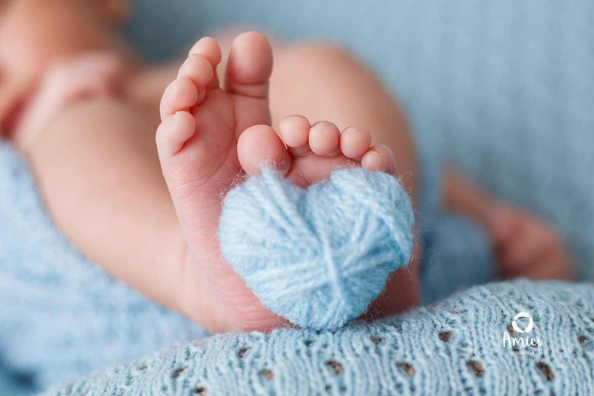 Fotografia macro, destacando detalhes dos pés do bebê (como os dedinhos e textura da pele) segurando um pequeno coração azul.