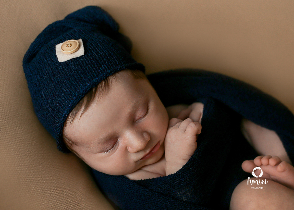 Bebê recém nascido envolvido em um wrap azul marinho com touca, na pose barriguinha para cima.