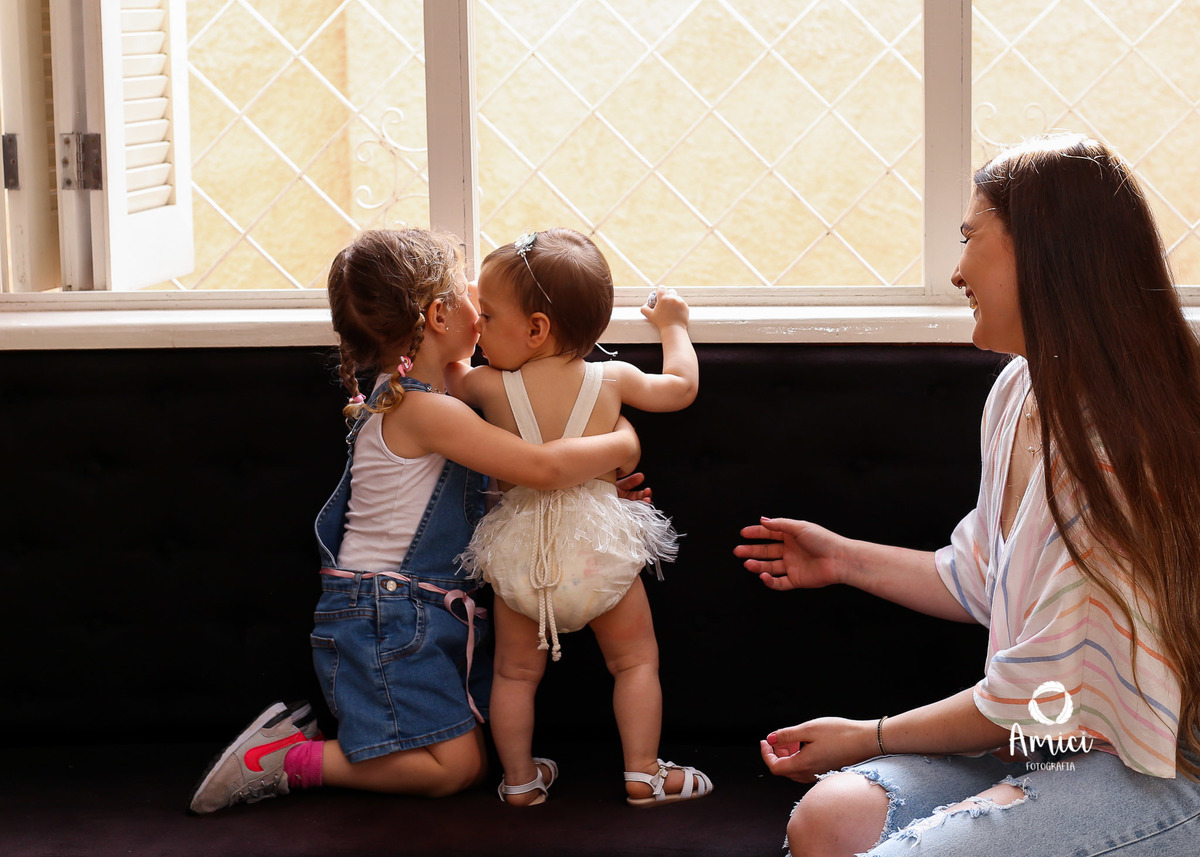 Fotografia infantil, primas se abraçando em frente à janela.