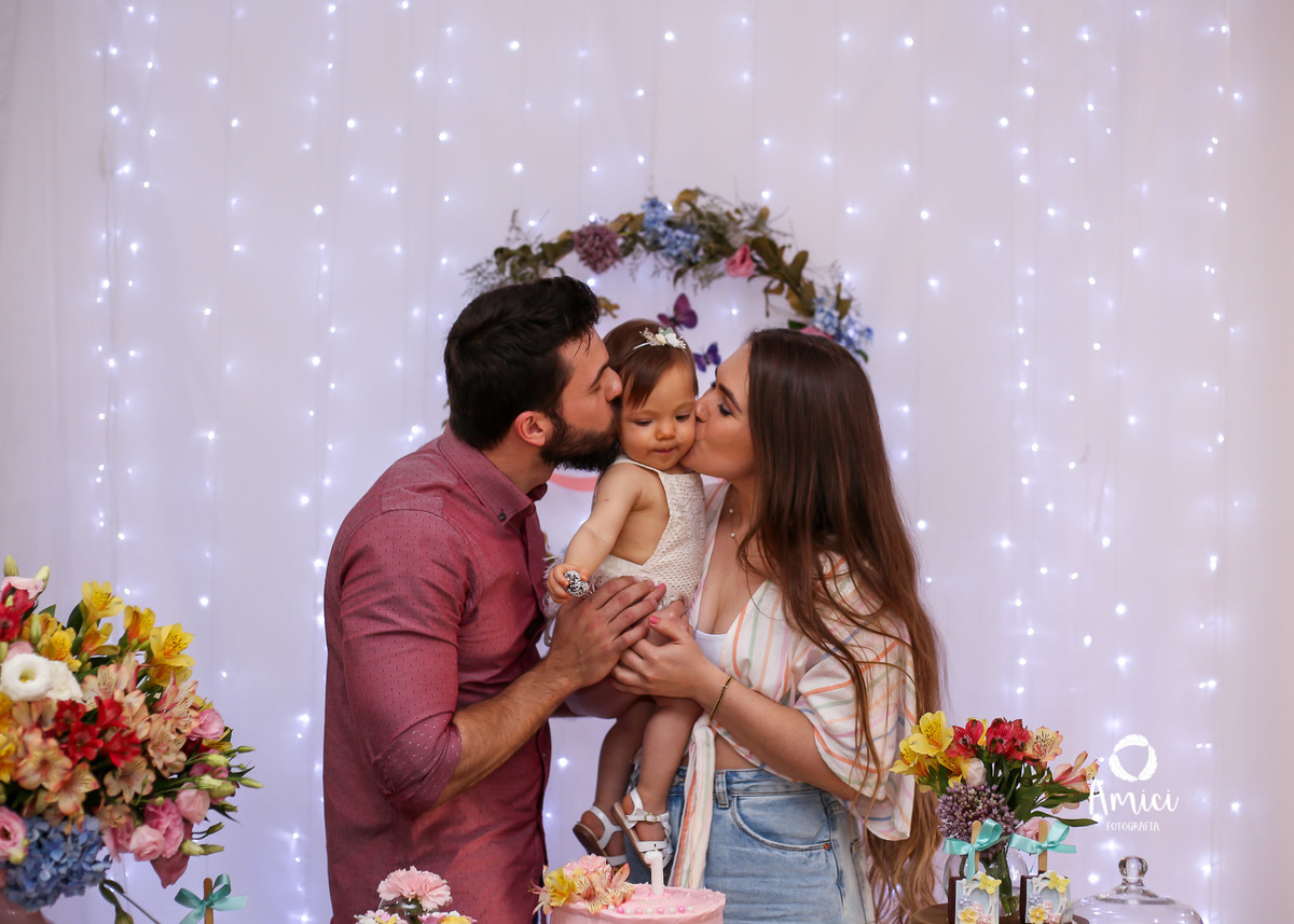 Festa infantil, mãe e pai dando beijo duplo no rosto da bebê.