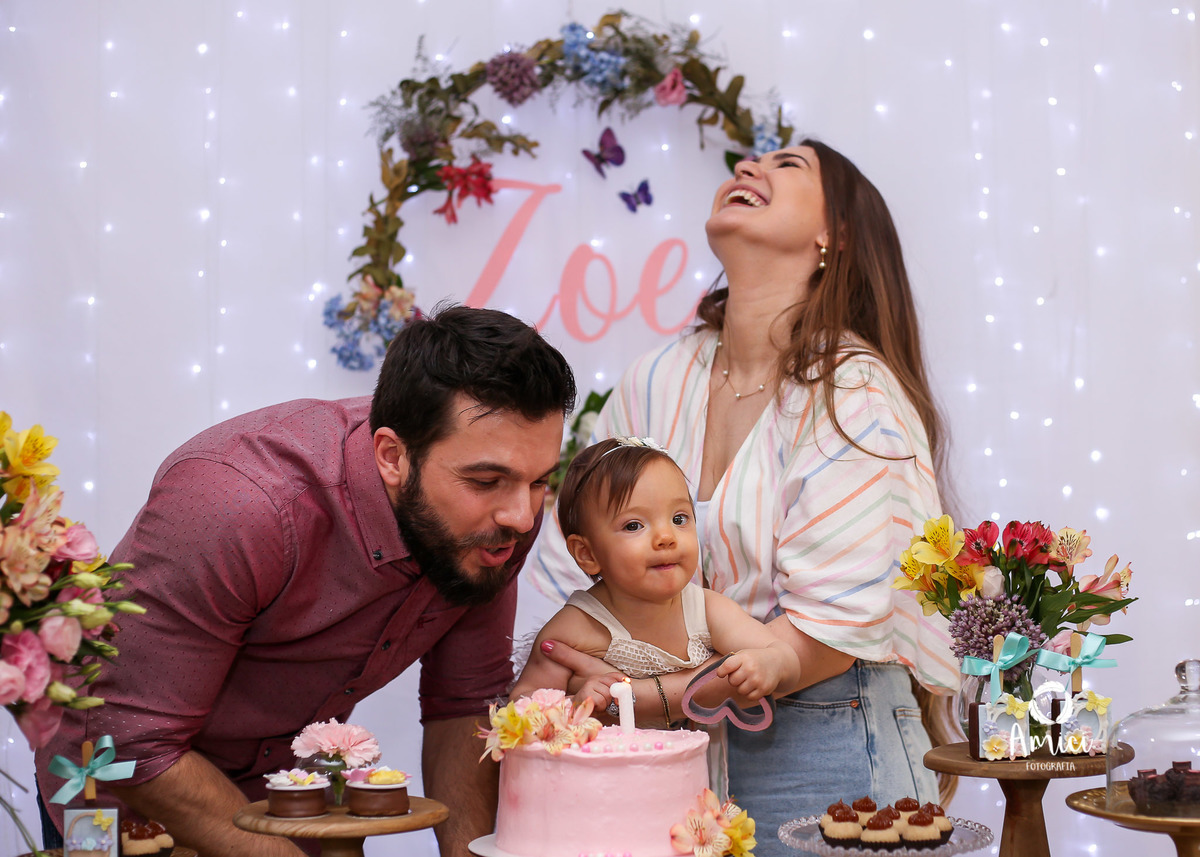 Evento infantil, pai assoprando a velinha, bebê fazendo careta e mãe gargalhando.