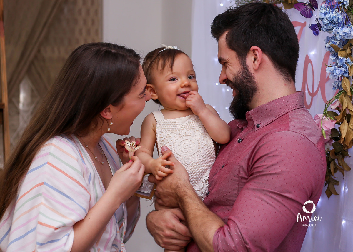 Fotografia infantil, pai e mãe brincando com a bebê que faz careta.