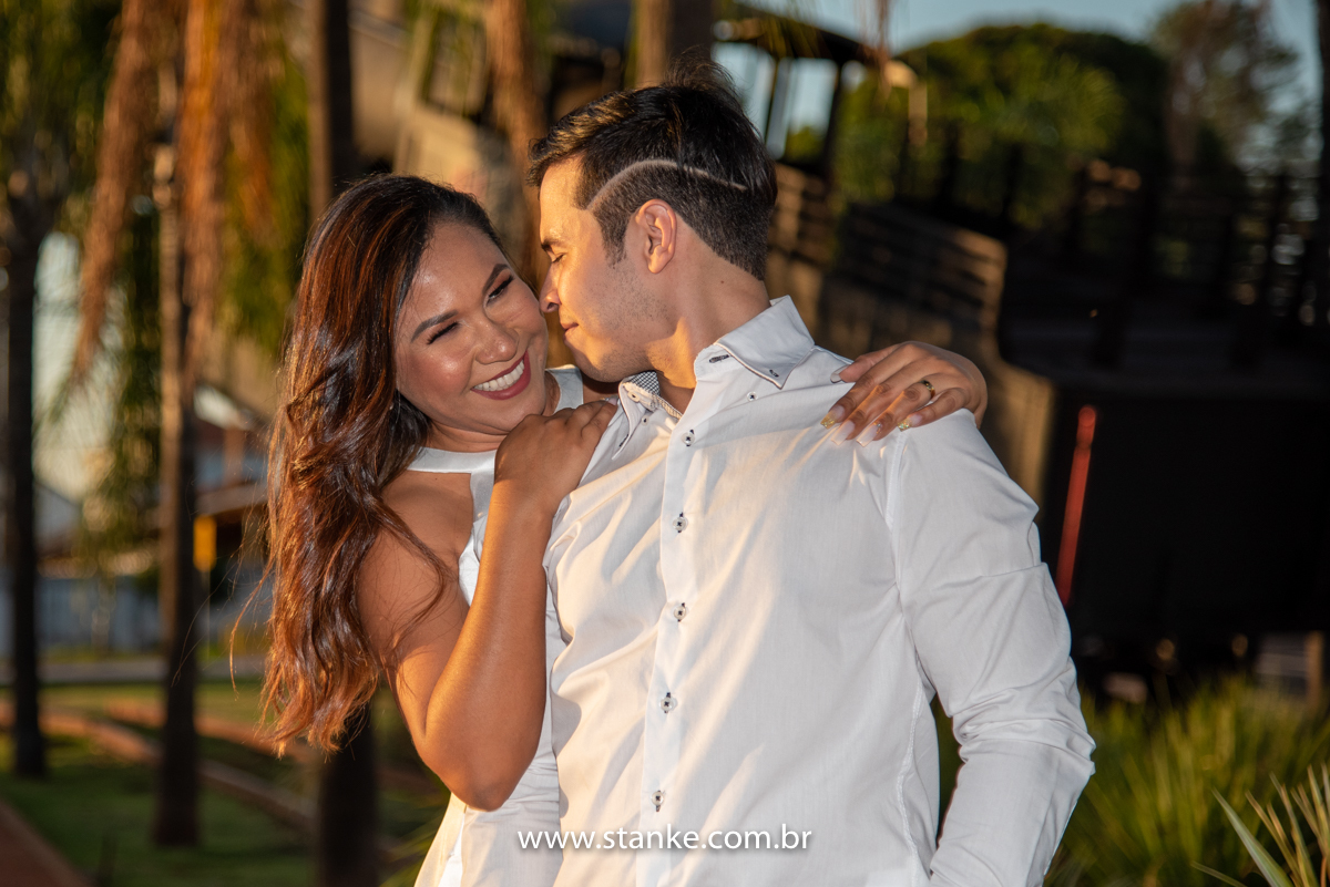 Ensaio pós casamento de Giseli e Aurélio, Maria Fumaça ao fundo e Giseli abraçando o ele por trás e com as mão no ombros dele e com sorriso e ele olhando para ela. Fotos feitas no Memorial Ferroviário, em Campo Grande-MS, por Pedro Stanke