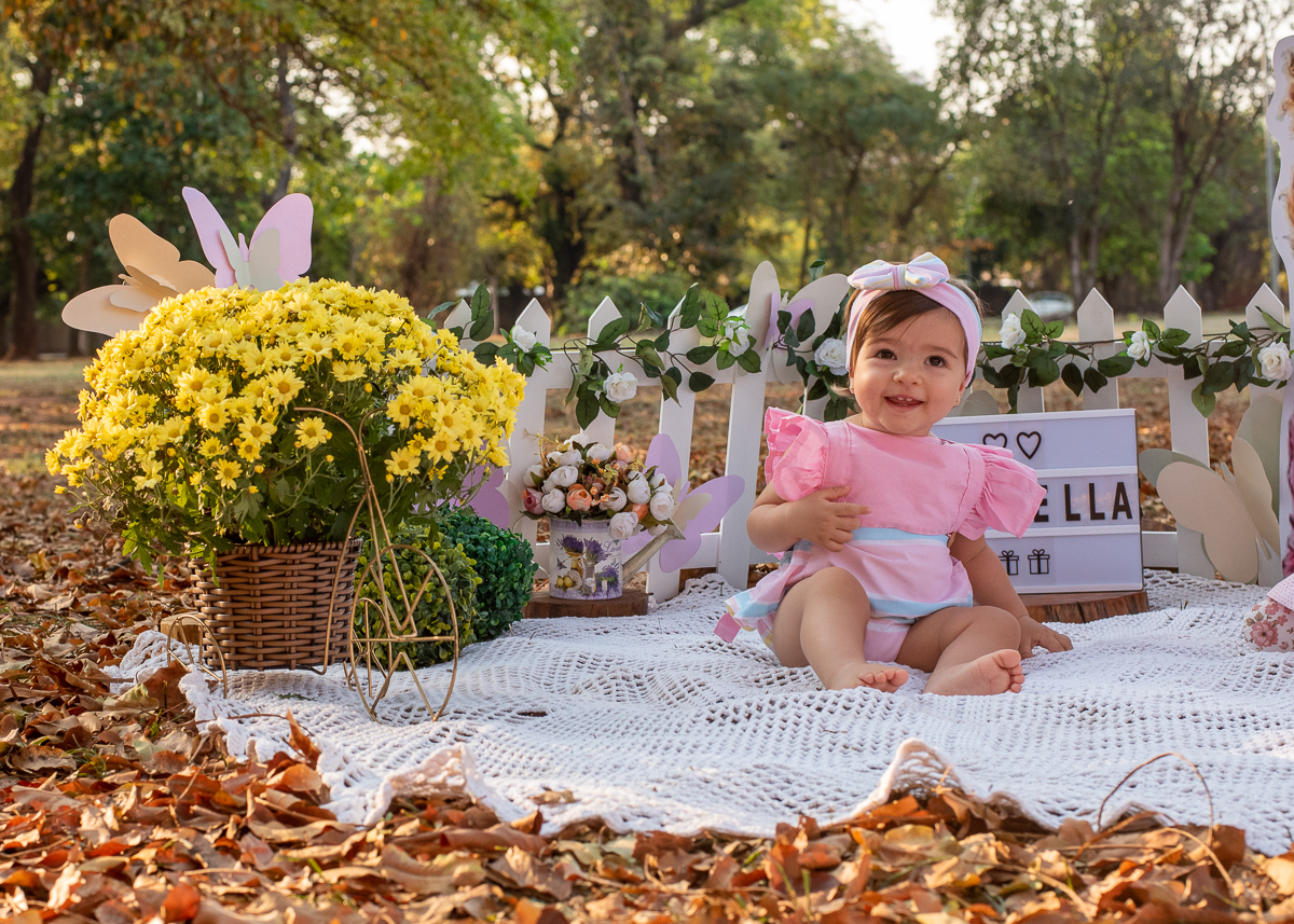 Smash the Cake da Gabriella, com a sua roupinha linda e sentadinha sobre um tapete de crochê e o chão cheio de folhas secas e em sua volta uma cerquinha de madeira e flores, Fotos feitas pelo fotógrafo Pedro Stanke em Campo Grande-MS. 