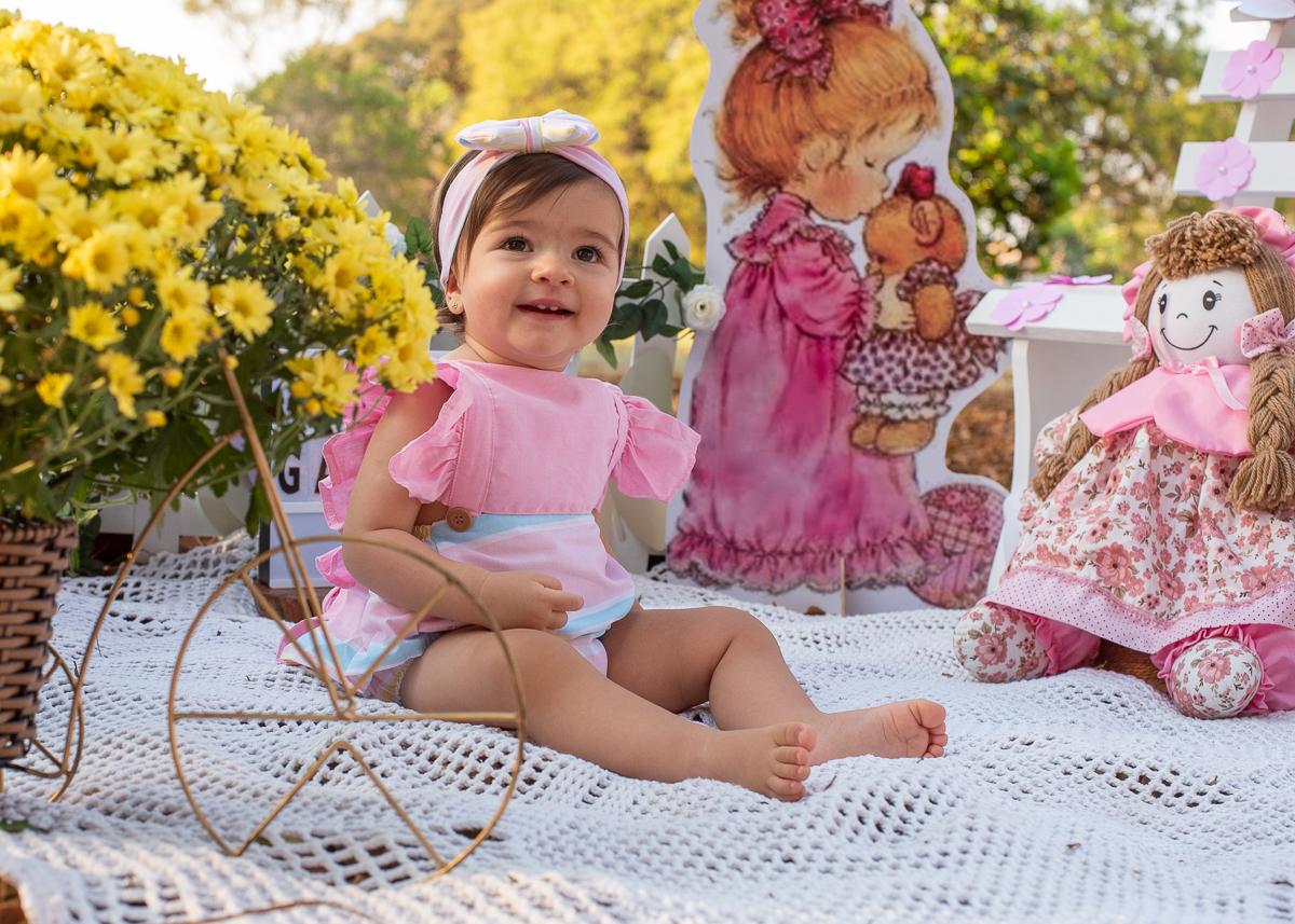 Smash the Cake da Gabriella, com lindo sorriso e sentadinha sobre um tapete de crochê e o chão cheio de folhas secas e em sua volta uma cerquinha de madeira e flores, Fotos feitas pelo fotógrafo Pedro Stanke em Campo Grande-MS. 