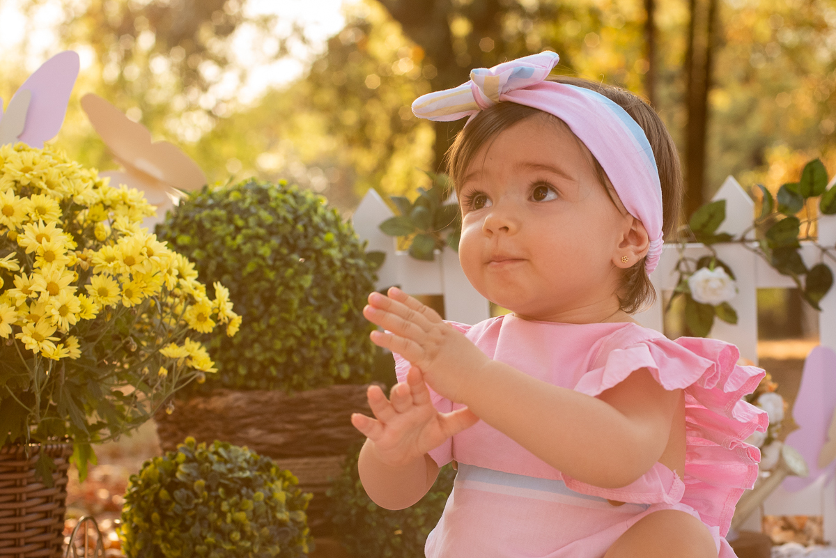 Smash the Cake da Gabriella, batendo palmas e sentadinha no chão e em sua volta uma cerquinha de madeira e flores, e fundo super iluminado pelo sol refletindo nas árvores. Fotos feitas pelo fotógrafo Pedro Stanke em Campo Grande-MS. 