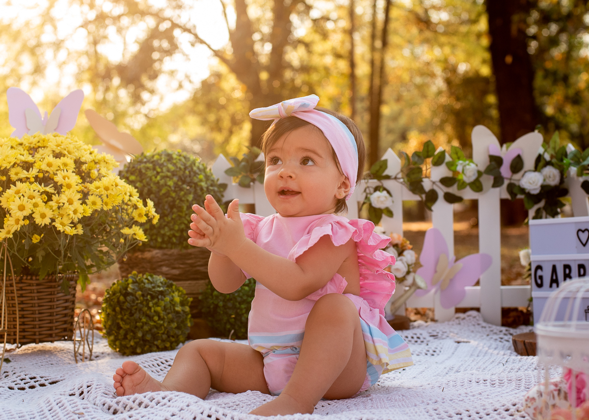 Smash the Cake da Gabriella, batendo palmas e sorrindo e sentadinha no chão e uma cerquinha de madeira e arranjos de flores atrás, e no fundo super iluminado pelo sol refletindo nas árvores. Fotos feitas pelo fotógrafo Pedro Stanke em Campo Grande-MS. 