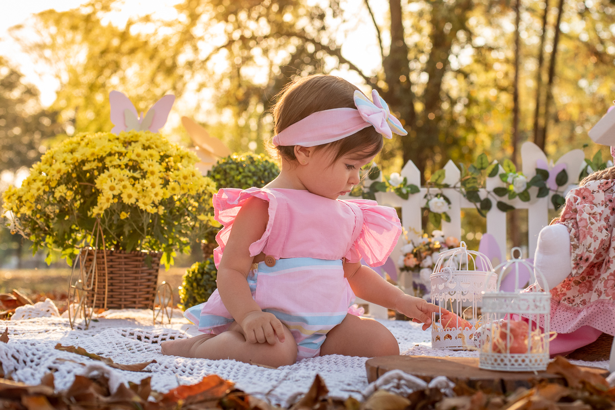 Smash the Cake da Gabriella, brincando com a decoração, com uma cerquinha de madeira e arranjos de flores atrás, e no fundo super iluminado pelo sol refletindo nas árvores. Fotos feitas pelo fotógrafo Pedro Stanke em Campo Grande-MS. 