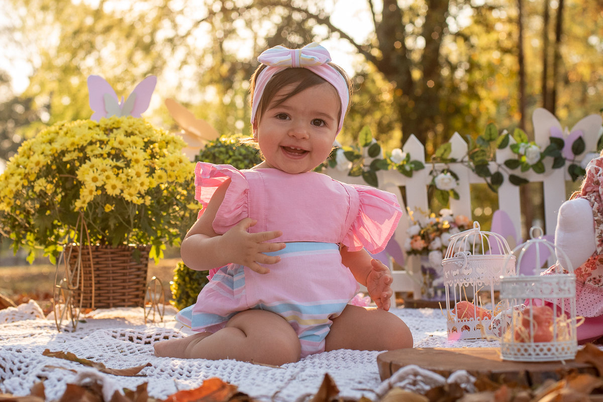 Smash the Cake da Gabriella, olhando para câmera com lindo sorriso, e com uma cerquinha de madeira e arranjos de flores atrás, e no fundo super iluminado pelo sol refletindo nas árvores. Fotos feitas pelo fotógrafo Pedro Stanke em Campo Grande-MS. 