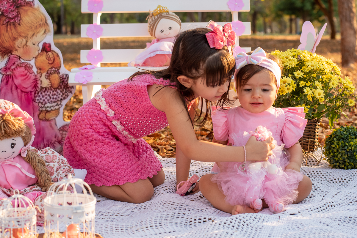 Smash the Cake da Gabriella e sua irmã brincando, com um banco branco e algumas bonecas e arranjos de flores atrás, e no fundo super iluminado pelo sol refletindo nas árvores. Fotos feitas pelo fotógrafo Pedro Stanke em Campo Grande-MS. 