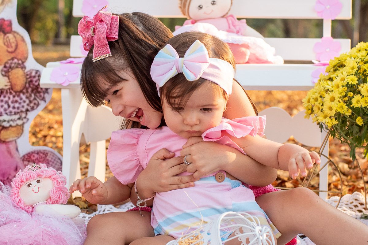 Smash the Cake da Gabriella sendo agarrada pela sua irmã, e um banco branco e algumas bonecas e arranjos de flores atrás, e o sol iluminando pela lateral e refletindo nas árvores. Fotos feitas pelo fotógrafo Pedro Stanke em Campo Grande-MS. 