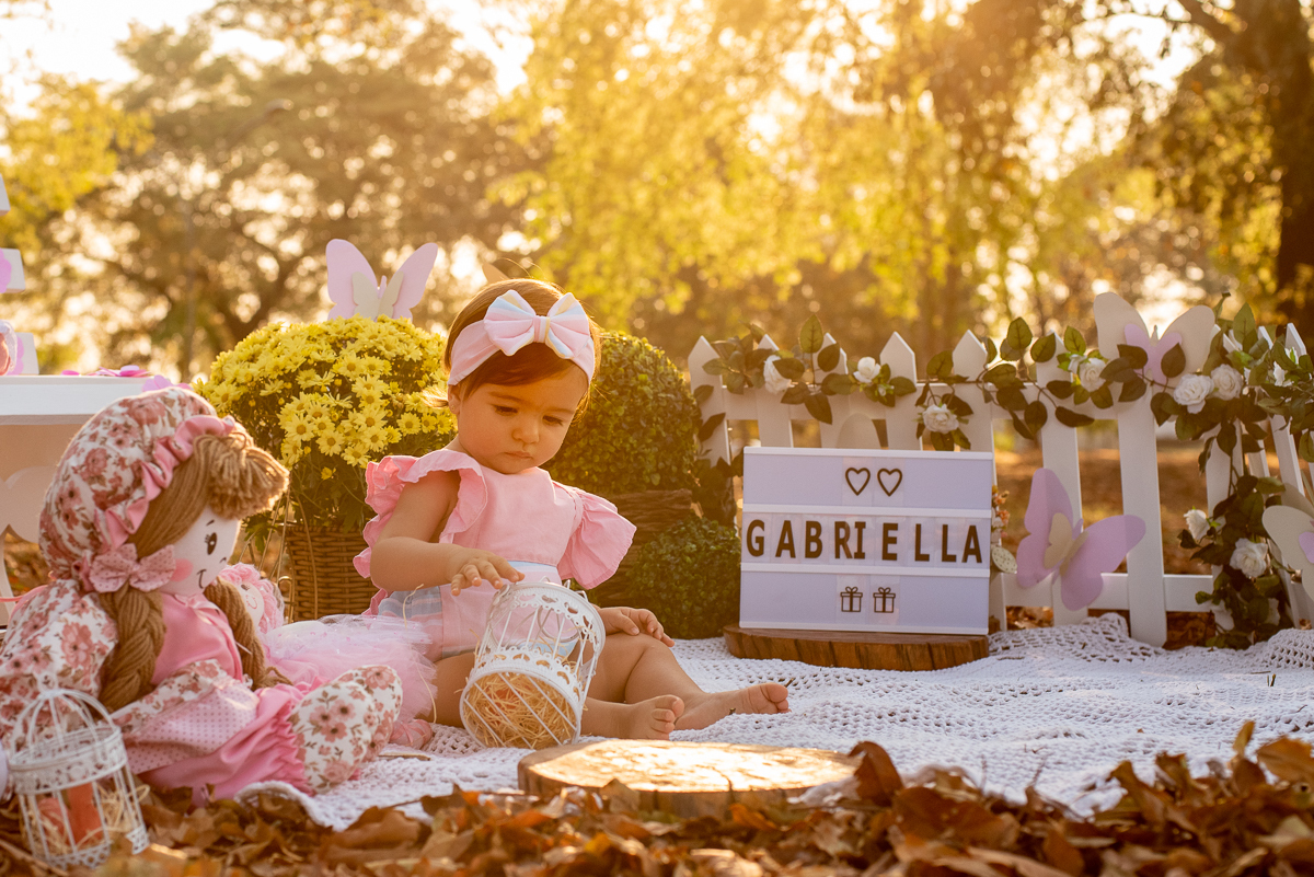 Smash the Cake da Gabriella brincando com a bonecas, com um banco branco de lado e algumas bonecas e arranjos de flores atrás, e no fundo super iluminado pelo sol refletindo nas árvores. Fotos feitas pelo fotógrafo Pedro Stanke em Campo Grande-MS. 