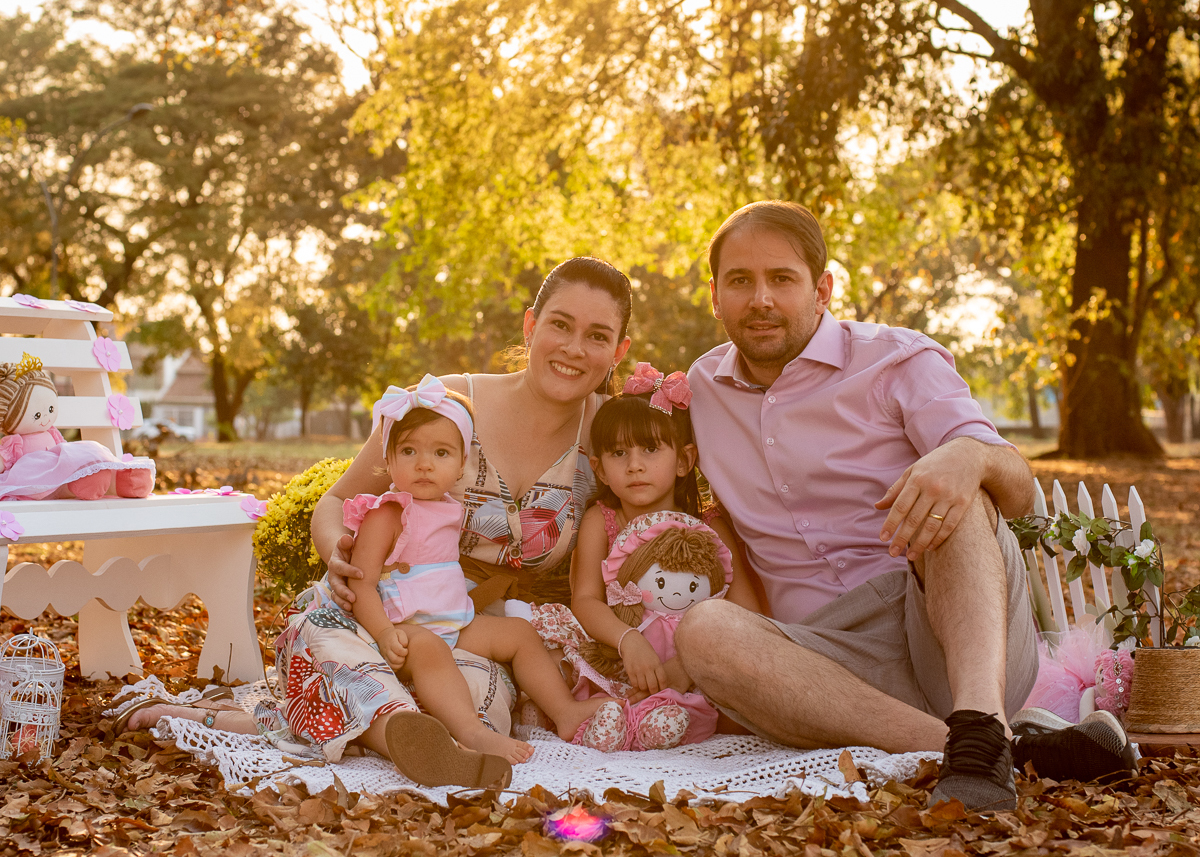 Smash the Cake da Gabriella e sua família todos posando para as fotos, com um banco branco e algumas bonecas e no fundo super iluminado pelo sol refletindo nas árvores. Fotos feitas pelo fotógrafo Pedro Stanke em Campo Grande-MS. 