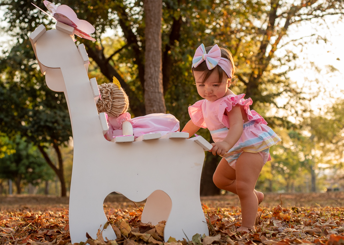 Smash the Cake da Gabriella, de pé e apoiando no banco branco e brincando com a boneca, e chão cheio de folhas secas, e no fundo super iluminado pelo sol refletindo nas árvores e nas folhas. Fotos feitas pelo fotógrafo Pedro Stanke em Campo Grande-MS. 