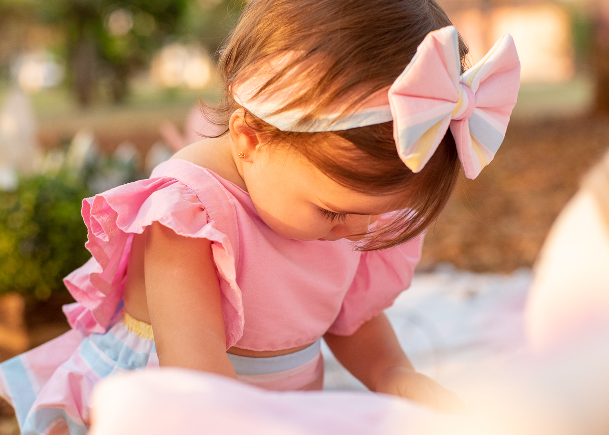 Smash the Cake da Gabriella, Um close da Gabi de pé e apoiando no banco branco e brincando com a boneca, e o sol iluminado o seu rosto e refletindo nas árvores e nas folhas. Fotos feitas pelo fotógrafo Pedro Stanke em Campo Grande-MS. 