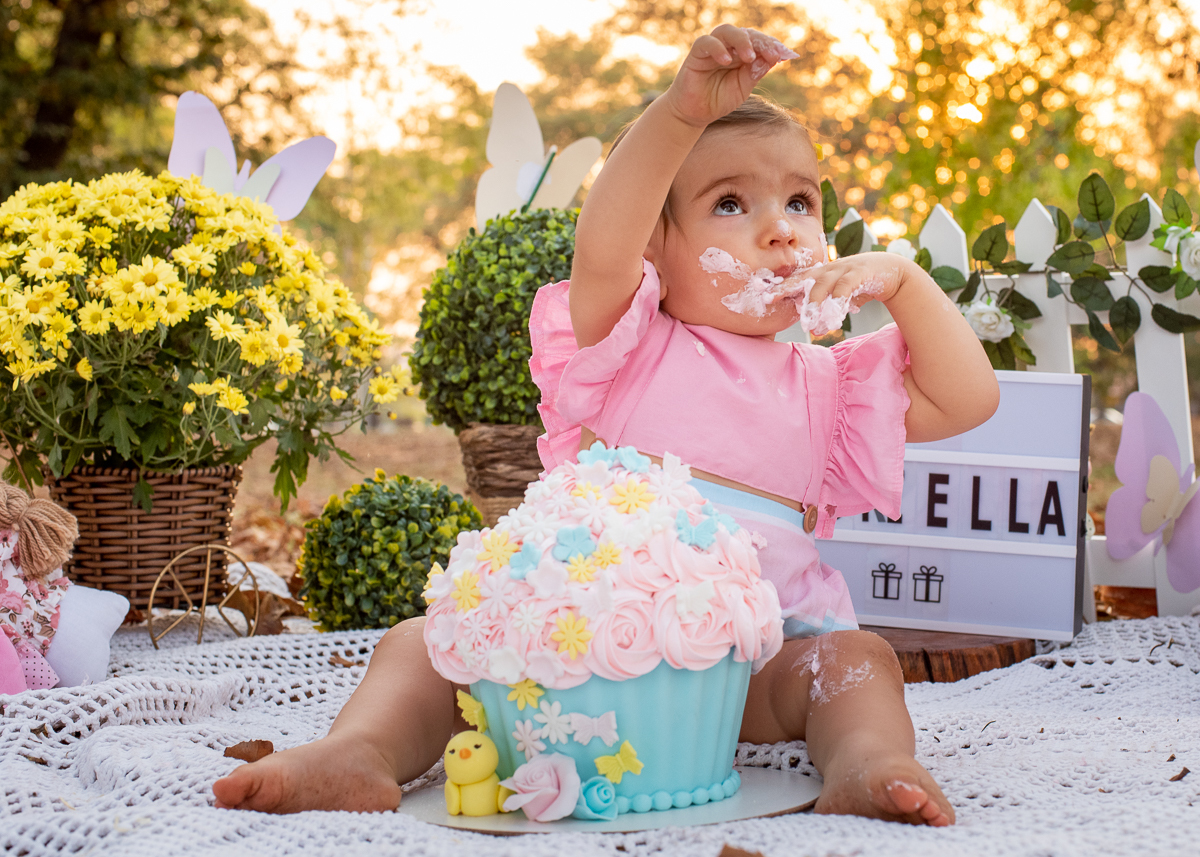Smash the Cake da Gabriella, a Gabi se lambuzando com o seu bolo, e no fundo uma cerquinha e arranjos de flores e o sol iluminando todo o fundo e repetindo nas árvores. Fotos feitas pelo fotógrafo Pedro Stanke em Campo Grande-MS. 