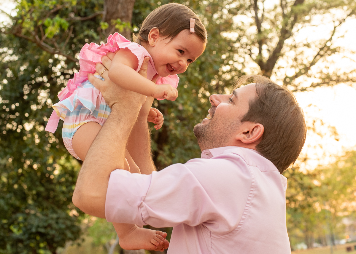 Smash the Cake da Gabriella, o papai a levantando para cima e a Gabi abrindo uma linda gargalhada e no fundo super iluminado pelo sol refletindo nas árvores. Fotos feitas pelo fotógrafo Pedro Stanke em Campo Grande-MS. 