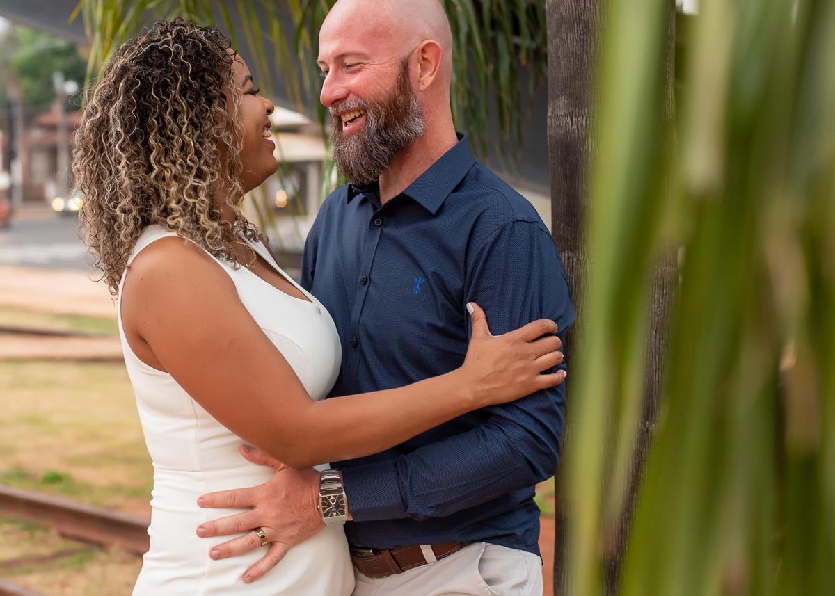 Ensaio de pós-casamento de Rosy e Alvaro, casal de frente um para o outro, ele encostado em uma palmeira e ela encostada nele, com algumas folhas no primeiro plano. Fotos feitas no Memorial Ferroviário, em Campo Grande-MS, pelo fotógrafo Pedro Stanke.