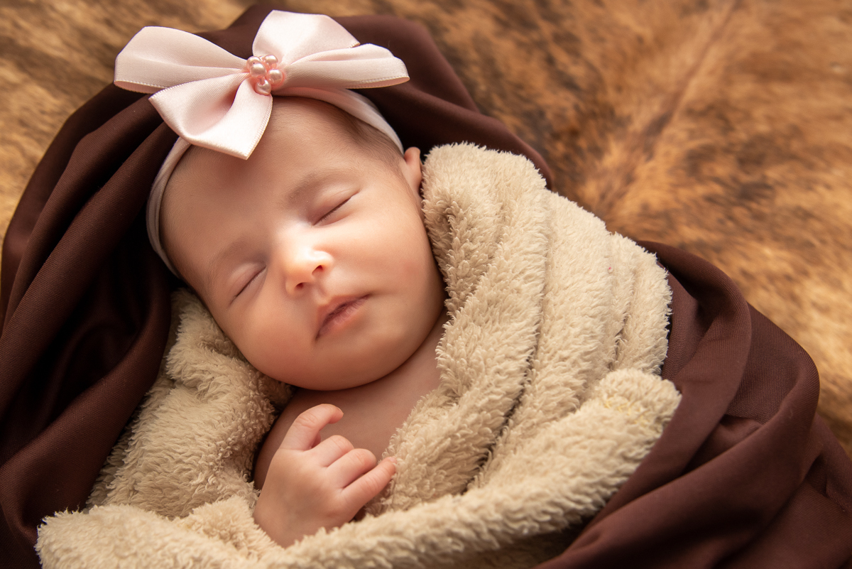 Ensaio newborn, Any com 25 dias. Any enrolada em seu cobertor creme e sobre ele o tecido marrom e no fundo o couro de boi de fundo. E ela com sua mãozinha solta,dormindo com sua tiara de laço dor de rosa. Fotos em Campo Grande-MS, feitas por Pedro Stanke.