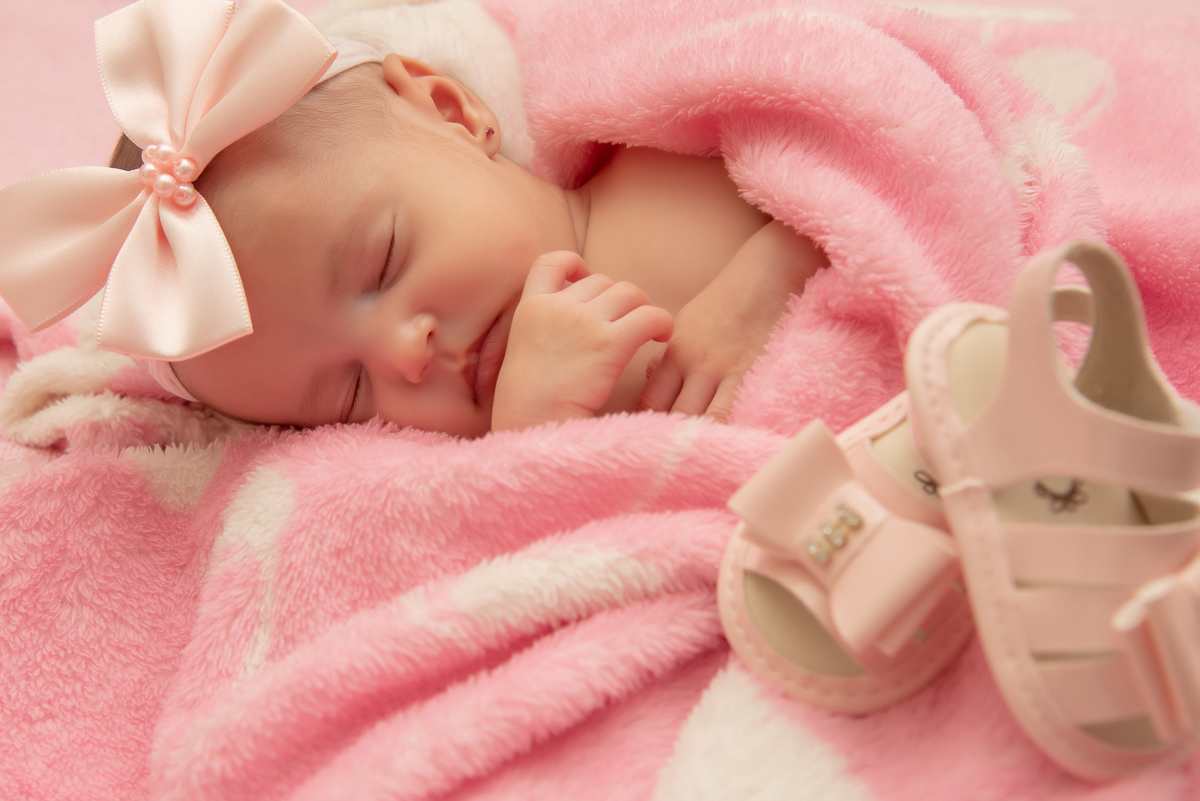 Ensaio newborn, Any com 25 dias. Any enrolada em seu cobertor cor de rosa. E ela com sua mãozinha no rosto, dormindo com sua tiara de laço dor de rosa. E os sapatinhos do papai de lado Fotos em Campo Grande-MS, feitas por Pedro Stanke.