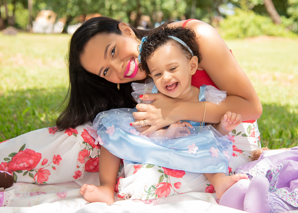 Ensaio Ana primeiro aninho, a mãe sentado no chão com a Ana no colo, com vestido azul com flores cor de rosa, e ambas sorrindo. Fotos feitas em Campo Grande-MS, por Pedro Stanke.