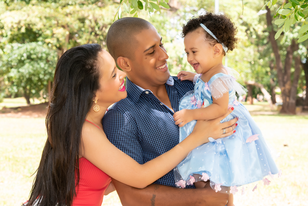 Ensaio Ana primeiro aninho, a Ana no colo do pai e a mãe ao lado do pai, e a Ana com lindo sorriso, com vestido azul com flores cor de rosa. Fotos feitas em Campo Grande-MS, por Pedro Stanke.