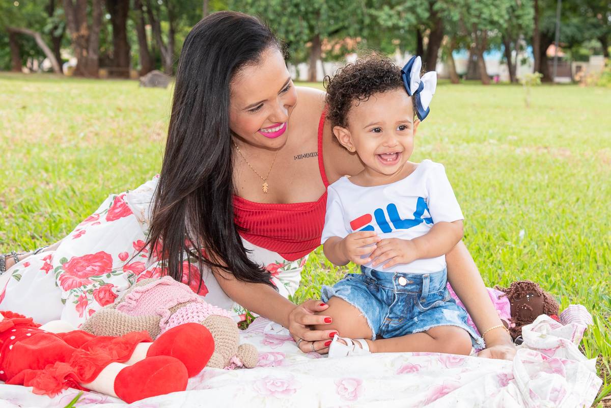 Ensaio Ana primeiro aninho. Ana sentada no chão com a sua mãe ao lado, com shorts jeans e camiseta branca, e lindo laço de cabelo azul e branco. Ambas sorrindo a vontade. Fotos feitas em Campo Grande-MS, por Pedro Stanke.
