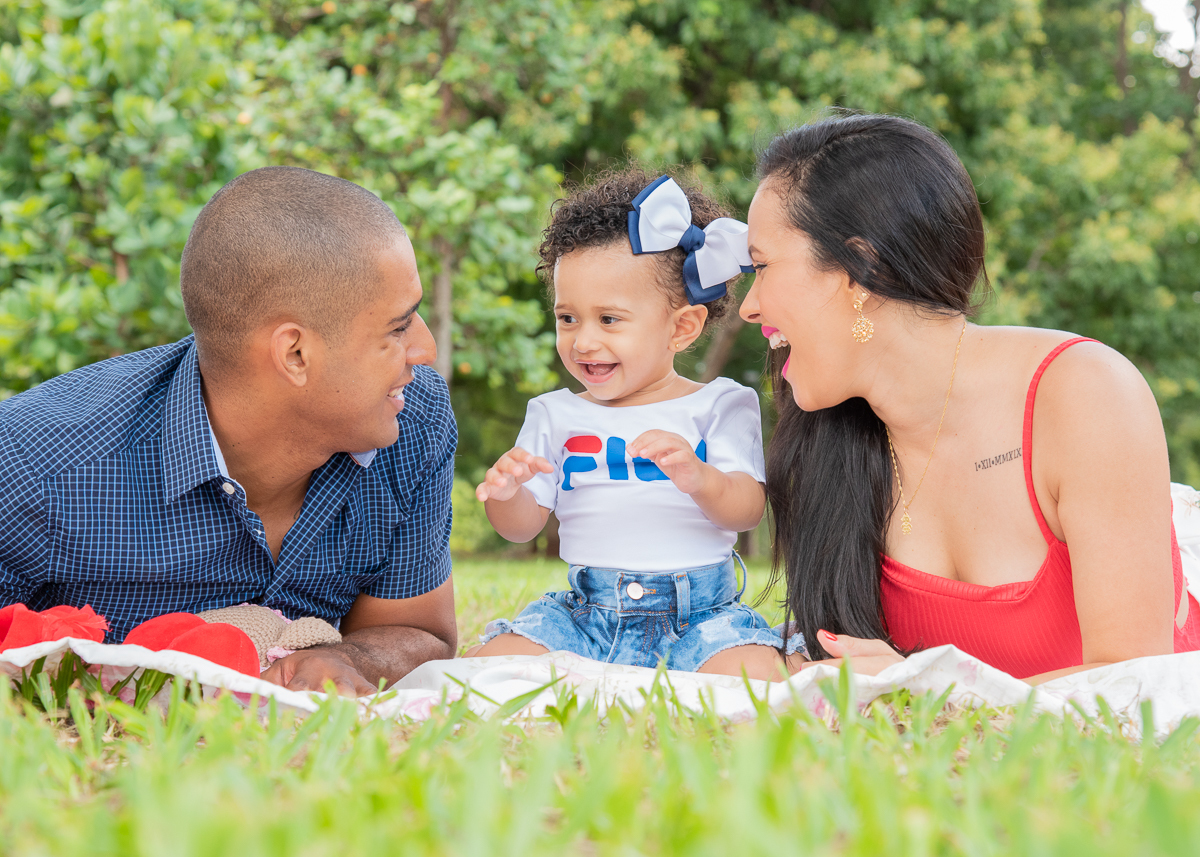 Ensaio Ana primeiro aninho. Ana sentada no chão com seus pais deitados ao lado dela, com shorts jeans e camiseta branca, e lindo laço de cabelo azul e branco. todos a vontade. Fotos feitas em Campo Grande-MS, por Pedro Stanke.