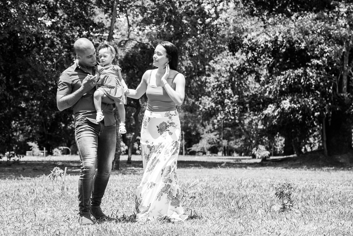 Foto preto e branco do Ensaio Ana primeiro aninho. Os pais caminhando com a Ana no colo do pai e mãe ao lado, ambos com olhares apaixonados pela Ana. Fotos feitas em Campo Grande-MS, por Pedro Stanke.