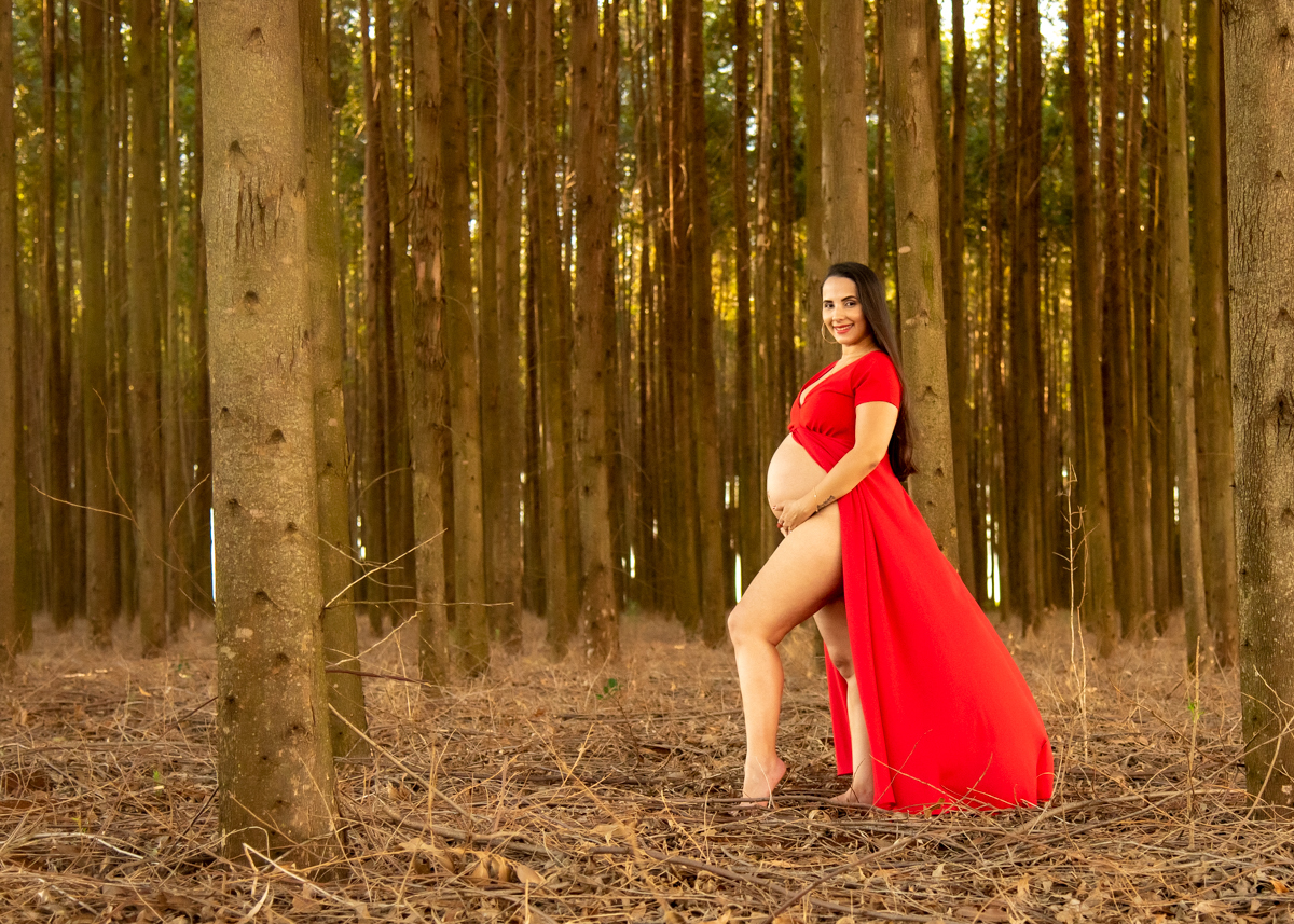 Ensaio gestante, Gestante com as mãos na barriga, com vestido vermelho, olhando para o fotógrafo, com lindo sorriso, no meio dos eucaliptos. Fotos feitas em Campo Grande-MS, por Pedro Stanke, fotógrafo de família e fotógrafo de gestantes.