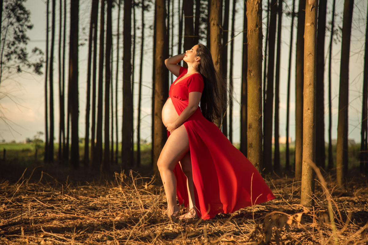 Ensaio gestante, Gestante com as mãos na barriga e nos cabelos, com vestido vermelho, olhando para cima, com os  eucaliptos no fundo. Fotos feitas em Campo Grande-MS, por Pedro Stanke, fotógrafo de família e fotógrafo de gestantes.