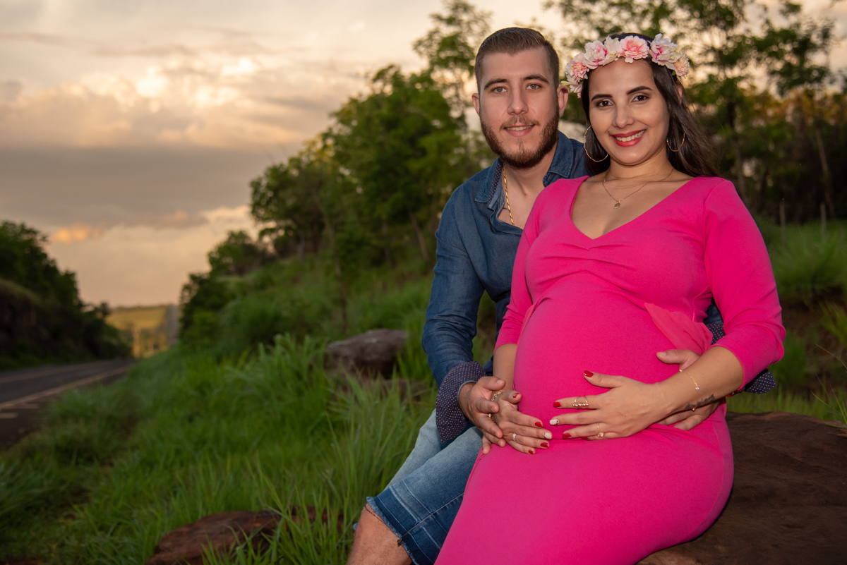 Ensaio gestante, casal sentados ao lado da rodovia, ele com camisa azul e ela com vestido rosa, ambos olhando para fotógrafo. Fotos feitas em Campo Grande-MS, por Pedro Stanke, fotógrafo de família e fotógrafo de gestantes.