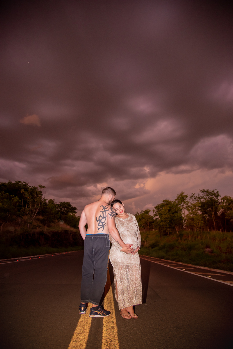 Ensaio gestante, casal abraçados de lado, ele sem camisa mostrando a tatu e ela com vestido dourado brilhante, no meio da rodovia, e nuvens de chuva enormes. Fotos feitas em Campo Grande-MS, por Pedro Stanke, fotógrafo de família e fotógrafo de gestantes.