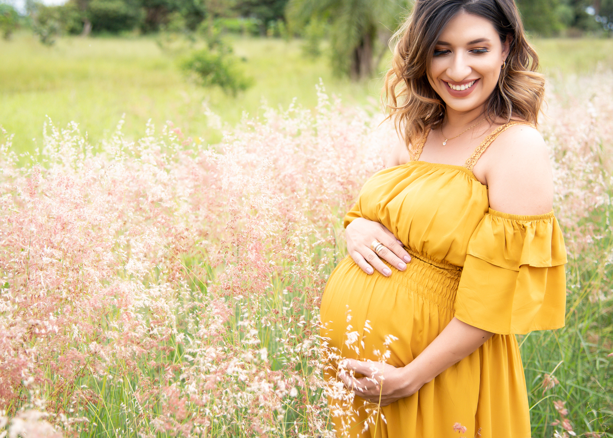 Ensaio Gestante, Donna e Allan. gestante posando com as mãos na barriga entre o capim alto com as pontas rosadas, com seu vestido longo cor mostarda, olhando de lado com lindo sorriso. Fotos feitas em Campo Grande-MS, por Pedro Stanke.