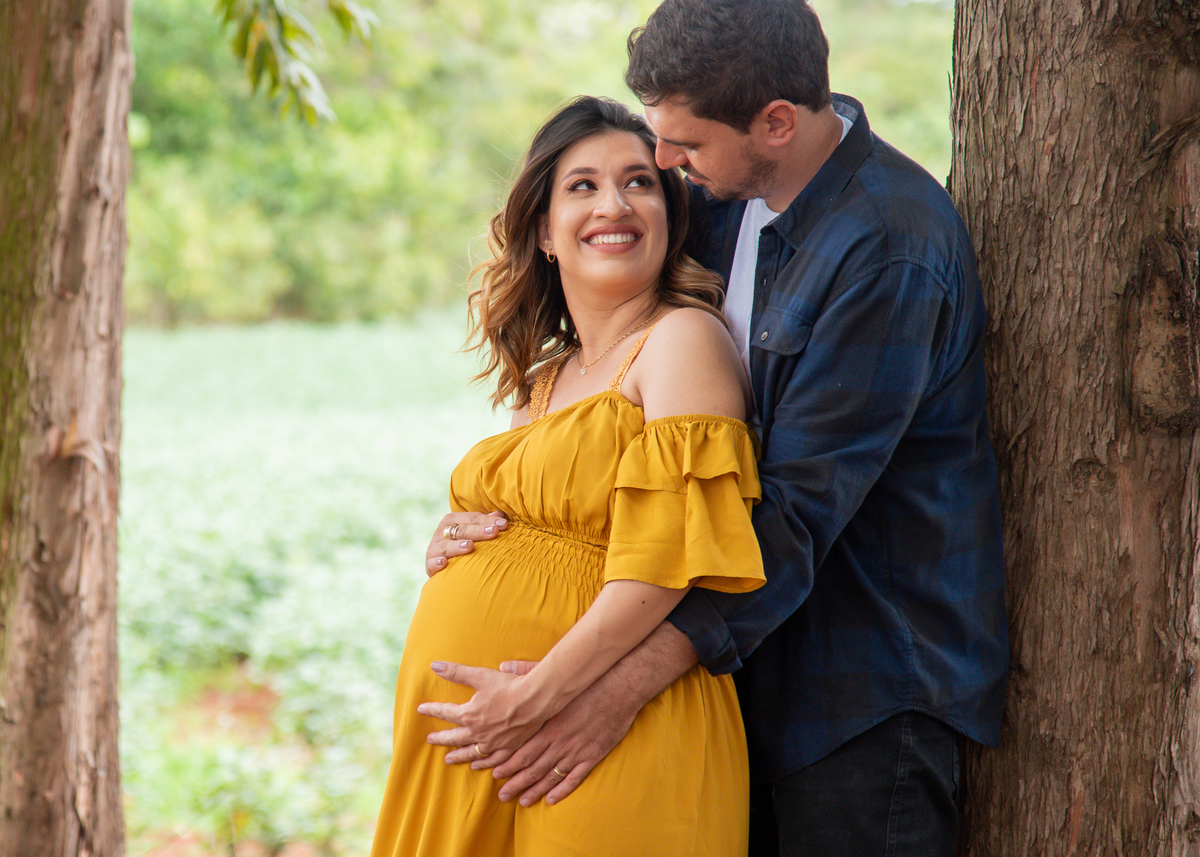 Ensaio Gestante, Donna e Allan, em uma estrada com eucaliptos. O pai abraçando a gestante por traz e com as mãos sobre a barriga da gestante, encostado na árvore e se olhando e sorrindo. Fotos feitas em Campo Grande-MS, por Pedro Stanke.