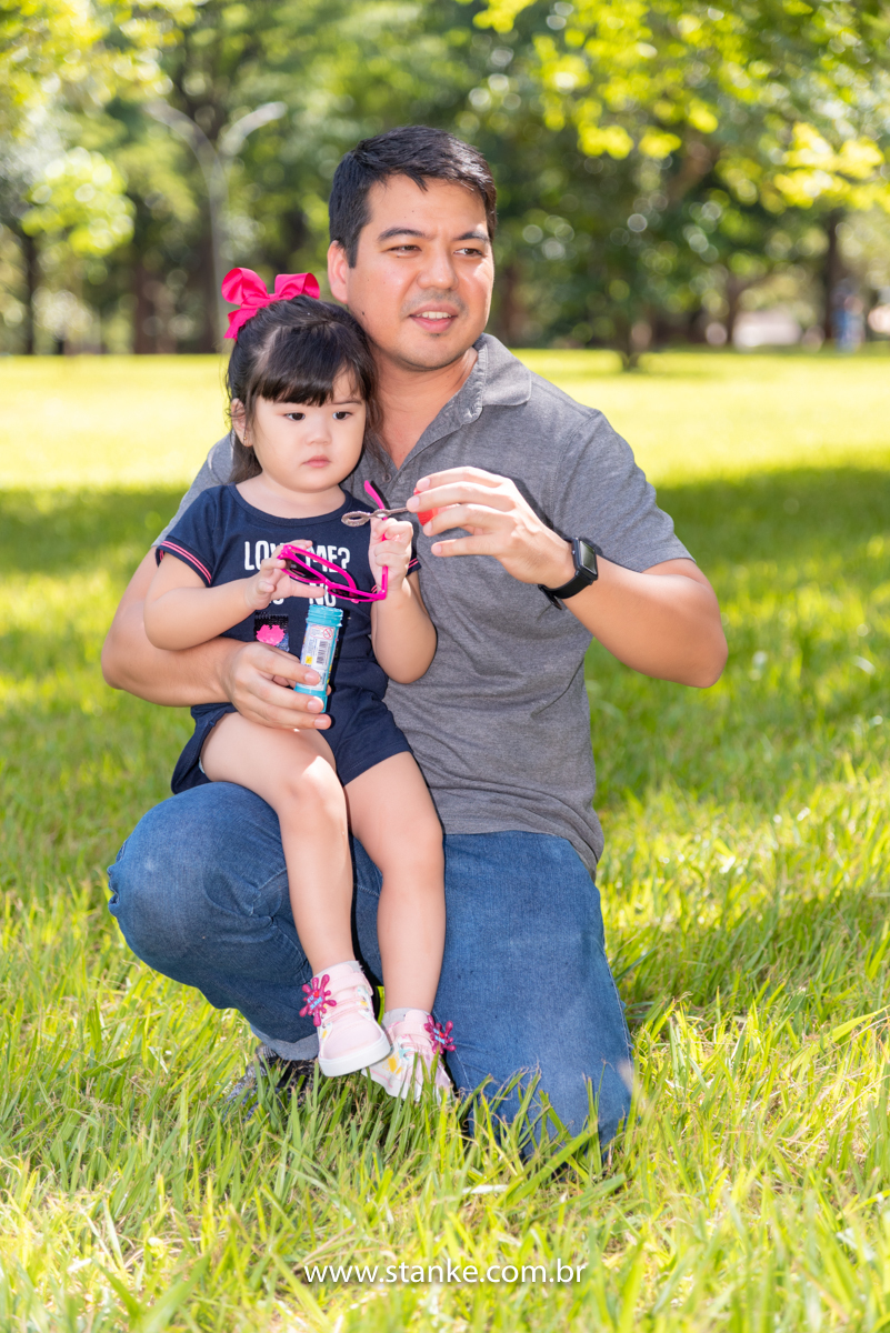 Ensaio Luísa de 2 anos, aniversariante com macacão azul e com linda fita vermelha prendendo os seus cabelos, muito séria e sentada no colo do seu pai, e ele fazendo bolhinhas de sabão. Fotos realizadas no Carandá Bosque, pelo fotógrafo Pedro Stanke. 