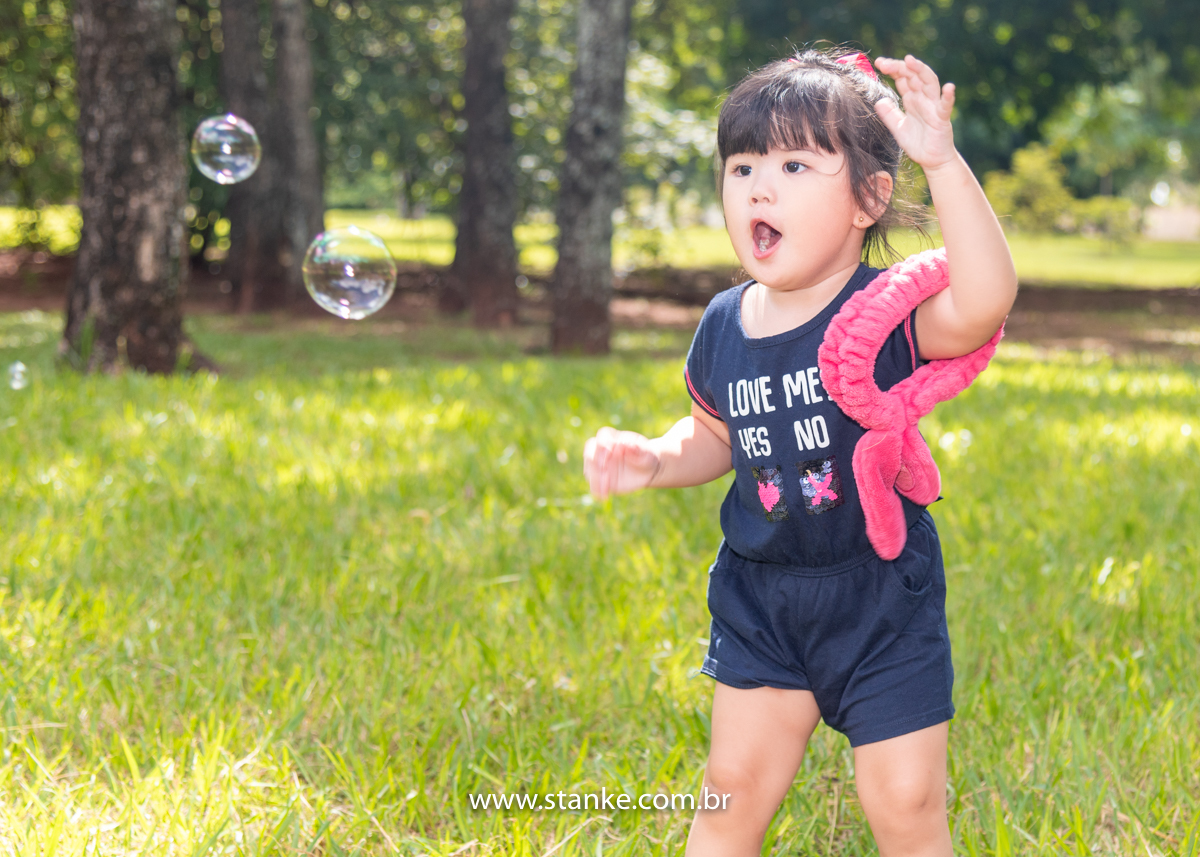 Ensaio Luísa de 2 anos, aniversariante com macacão azul e com linda fita vermelha prendendo os seus cabelos, brincado com a bolhinhas de sabão, e em seu braço o seu coelhinho de braço. Fotos realizadas no Carandá Bosque, pelo fotógrafo Pedro Stanke. 