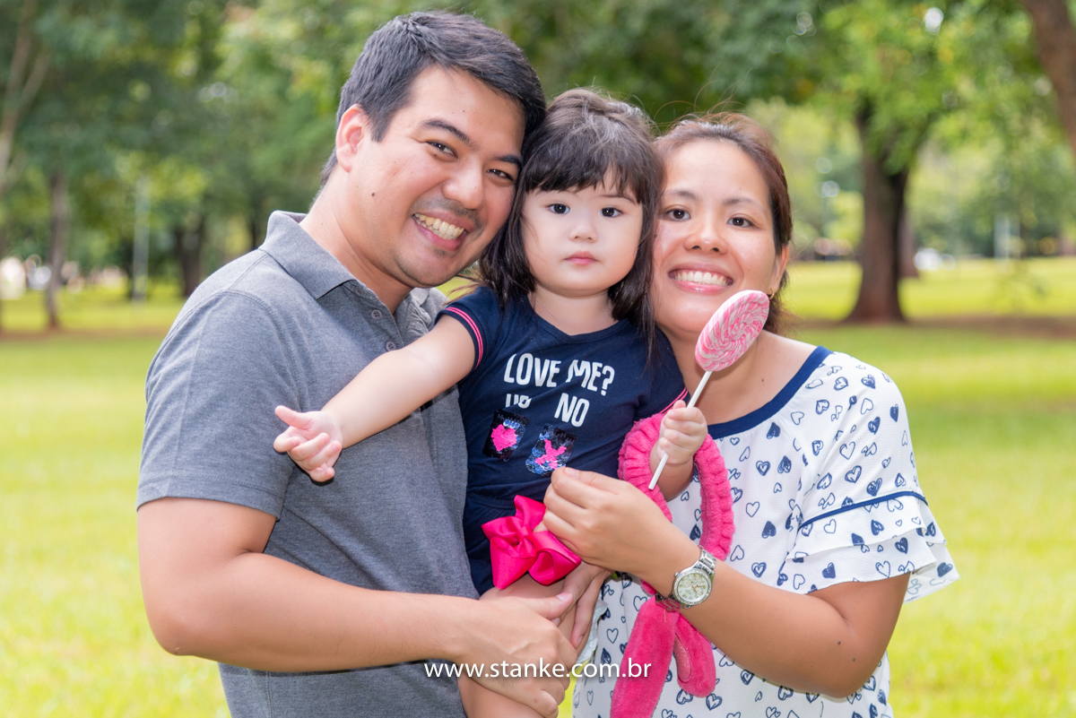 Ensaio Luísa de 2 anos, aniversariante entre seus pais e no colo do seu pai e ela séria, em seus braços o seu coelhinho e com seu pirolito colorido. Fotos realizadas no Carandá Bosque, pelo fotógrafo Pedro Stanke. 