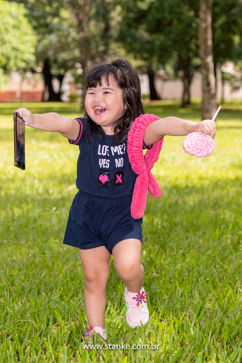 Ensaio Luísa de 2 anos, aniversariante com macacão azul, correndo de braços abertos e segurando celular, enorme pirolito colorido e em seu braço o seu coelhinho de braço. E lindo sorriso. Fotos realizadas no Carandá Bosque, pelo fotógrafo Pedro Stanke. 