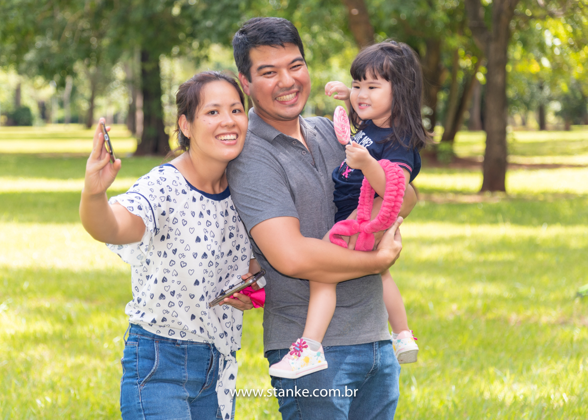 Ensaio Luísa de 2 anos, aniversariante no colo seu pai e sua mãe fazendo uma foto com celular e ela com lindo sorriso, em seus braços o seu coelhinho e com seu pirolito colorido. Fotos realizadas no Carandá Bosque, pelo fotógrafo Pedro Stanke. 