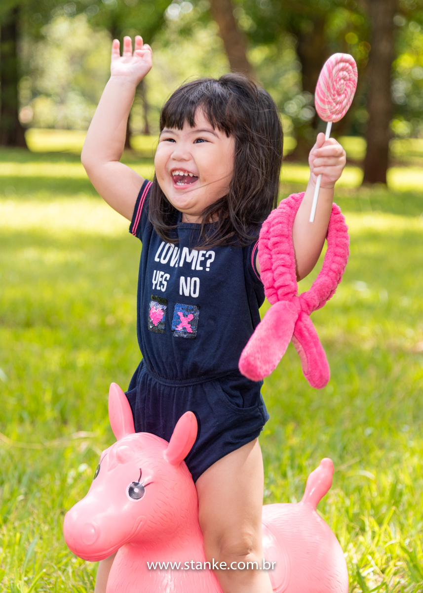 Ensaio Luísa de 2 anos, aniversariante com macacão azul, sentada em seu burrinho rosa, de braços abertos, enorme pirolito colorido e em seu braço o seu coelhinho de braço. E lindo sorriso. Fotos realizadas no Carandá Bosque, pelo fotógrafo Pedro Stanke. 