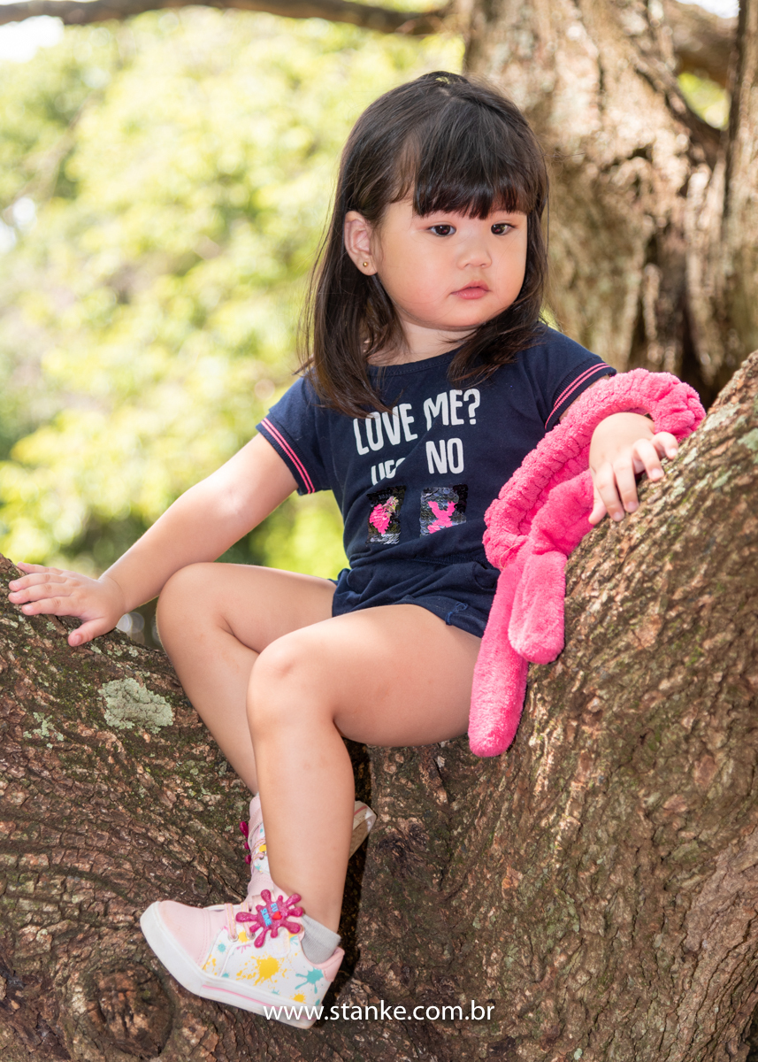 Ensaio Luísa de 2 anos, aniversariante com macacão azul, sentada nos galhos da árvore, sentada de lado, séria e com seu coelhinho de braço. Fotos realizadas no Carandá Bosque, pelo fotógrafo Pedro Stanke. 