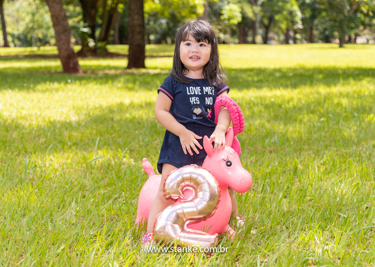 Ensaio Luísa de 2 anos, aniversariante com macacão azul, sobre o seu burrinho rosa, com o seu coelhinho no braço e um balão no formato do úmero 2 na frente. Fotos realizadas no Carandá Bosque, pelo fotógrafo Pedro Stanke. 