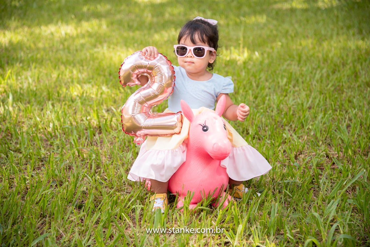 Ensaio infantil da Clara com 2 anos, Aniversariante, com seu vestido colorido e com seus óculos escuros rosa, sentada em um burro rosa e segurando um balão no formato número 2. Fotos feitas no Bosque da Paz, pelo fotógrafo Pedro Stanke. 